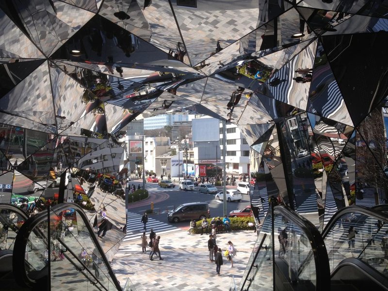 Building exit covered in mirror faces in all directions, reflecting the street outside, with a road crossing and pedestrians.jpg